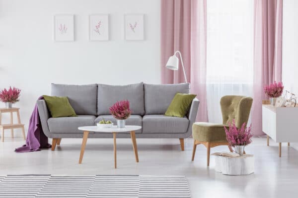 Grey settee with green cushions and purple blanket in real photo of white living room interior with coffee table with fruits and heather, posters on wall and window with white and dirty pink curtains